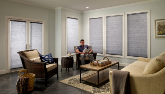 A living room with a man sitting on a gray armchair, reading a magazine with roller shades closed behind him.