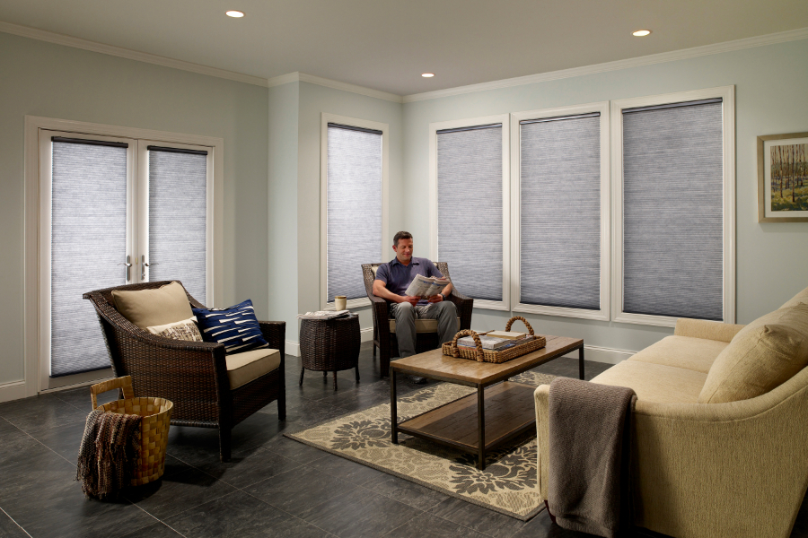 A living room with a man sitting on a gray armchair, reading a magazine with roller shades closed behind him.