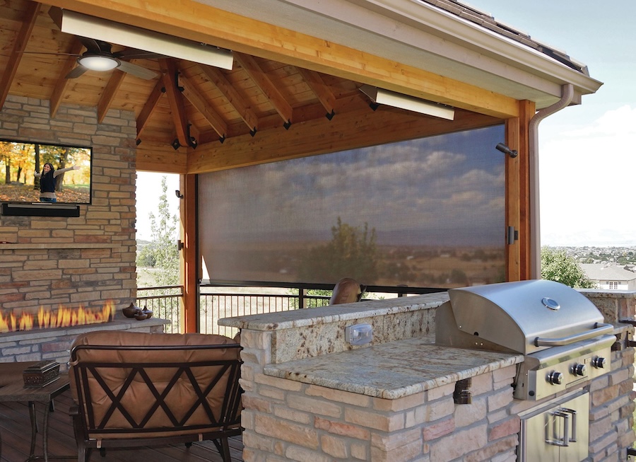 Outdoor grilling and seating area with a wall-mounted TV display and Insolroll solar shades partially lowered.
