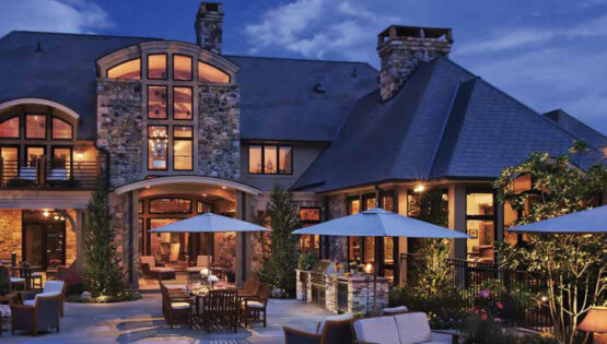 Outdoor view of a two-story home illuminated at dusk. Seating and umbrellas are in the foreground.