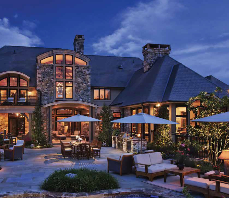 Outdoor view of a two-story home illuminated at dusk. Seating and umbrellas are in the foreground.