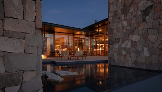 Outdoor view of a warmly lit desert home. A pool area in the foreground leads to a patio with wood chairs.